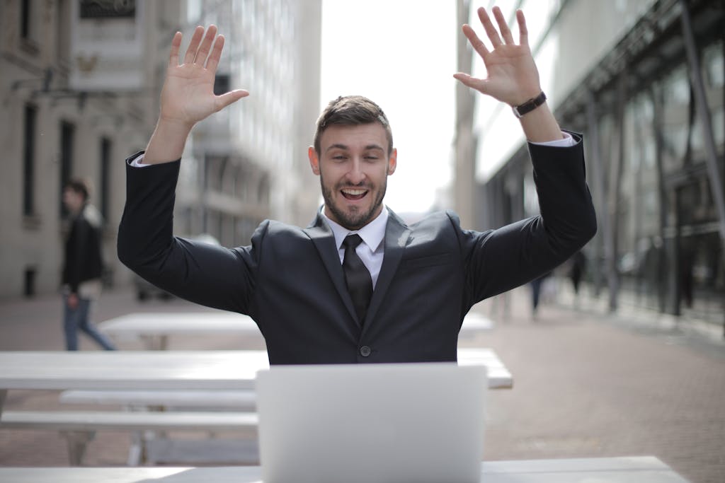 A joyful businessman raises his hands in excitement while working on a laptop outdoors.