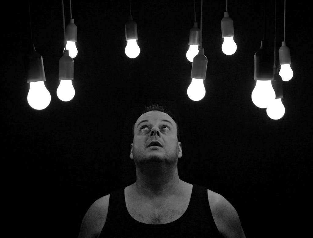 A man looks up thoughtfully at multiple illuminated light bulbs on a dark background.