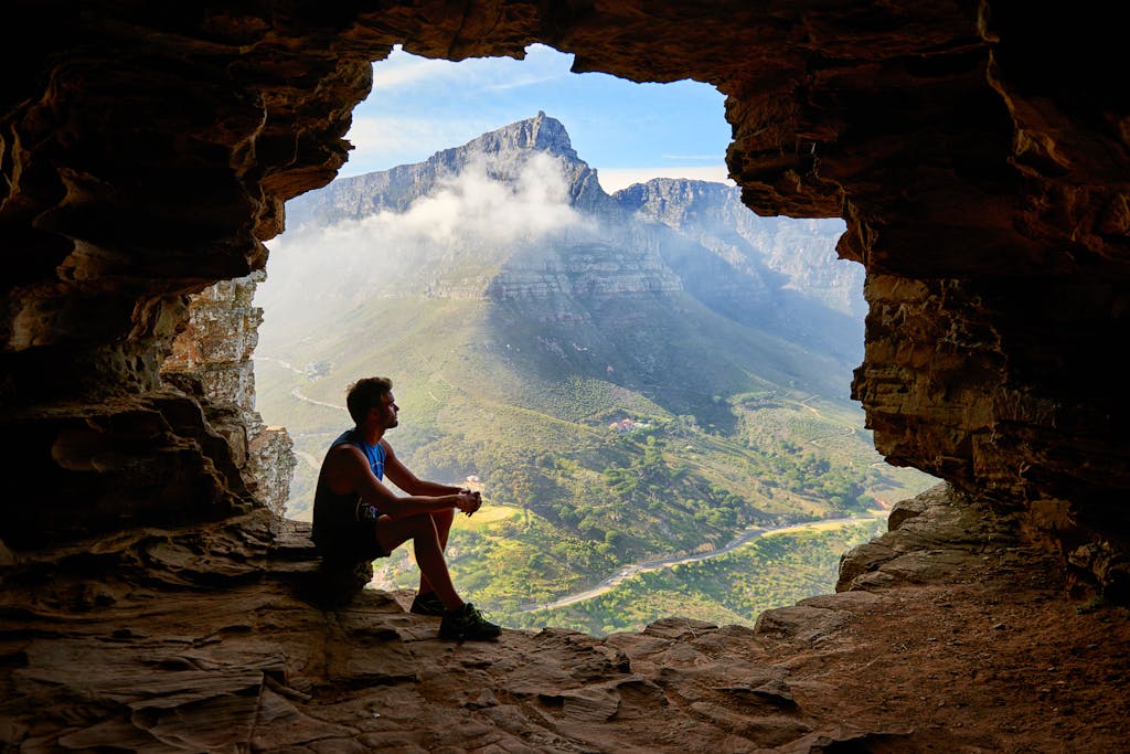 A man sitting in a cave overlooking a majestic mountain landscape under daylight.