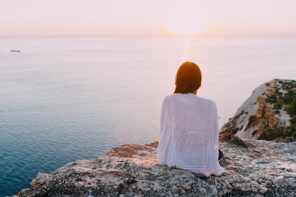 A serene view of a woman sitting on a rocky cliff, gazing at the ocean during sunset. Perfect for relaxation and travel themes.