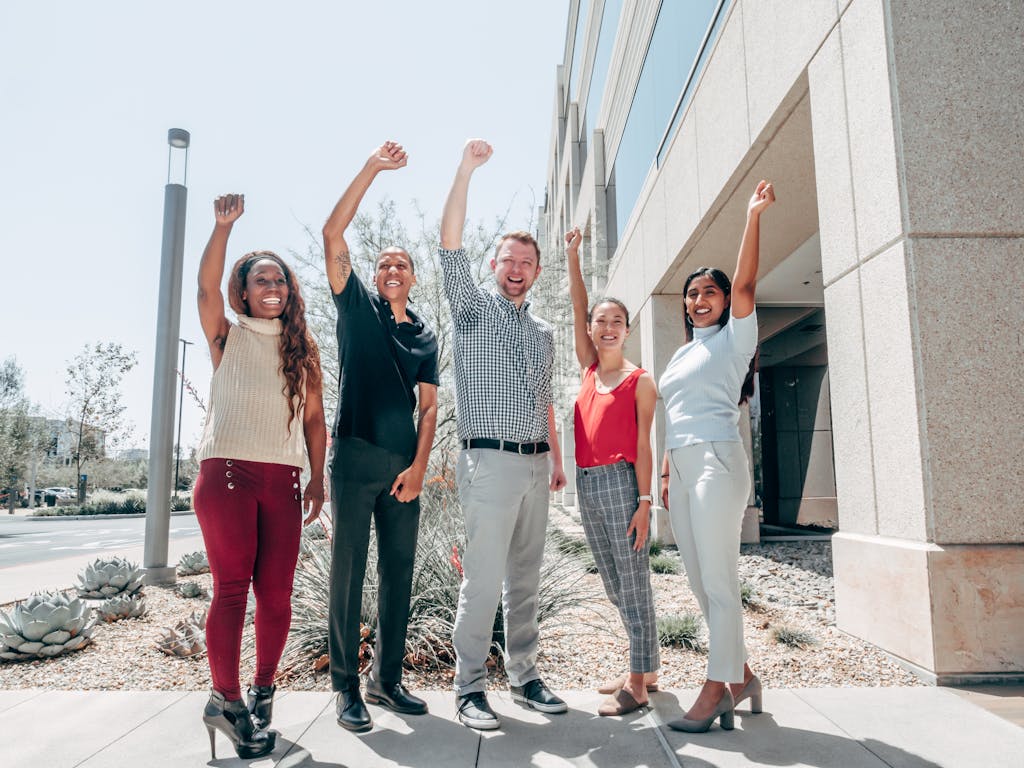 A group of diverse colleagues celebrate success with raised hands outside a modern building.