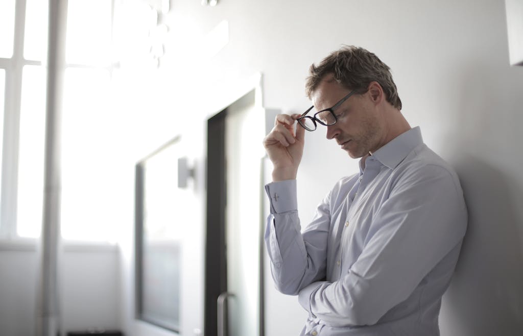 Thoughtful man in a bright room holding his glasses while leaning against a wall.