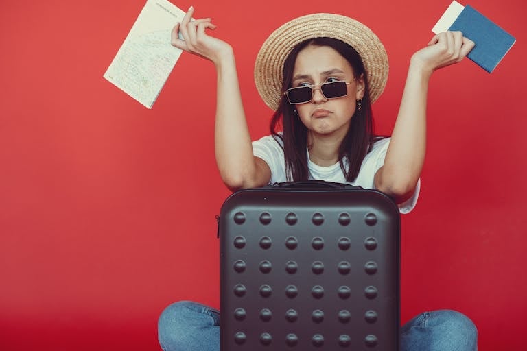 Young woman frustrated with travel plans, seated with suitcase and maps against a red background.