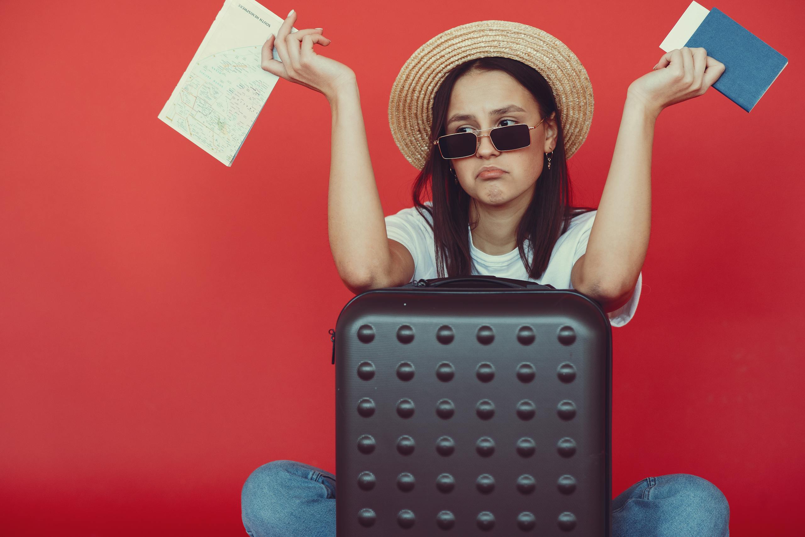 Young woman frustrated with travel plans, seated with suitcase and maps against a red background.